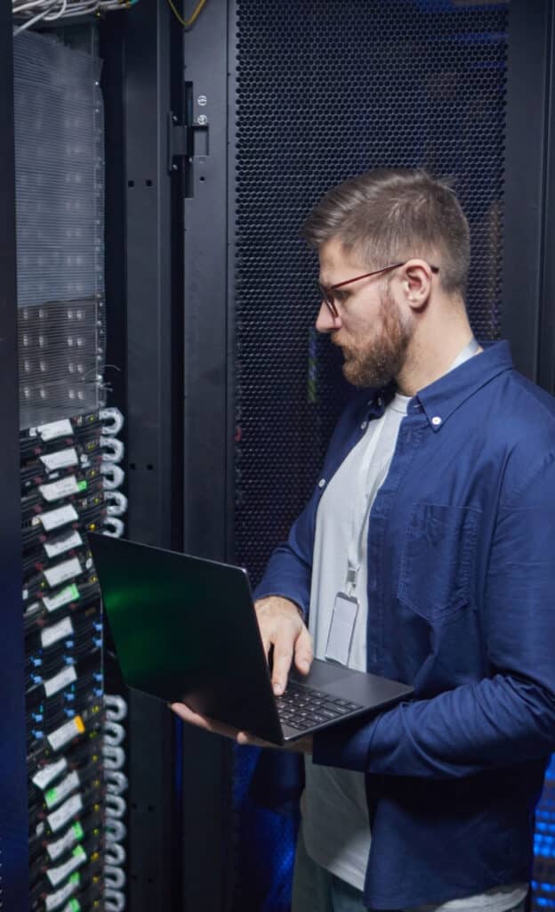 Wide shot of a technician performing maintenance in a switch room. Rows of black server cabinets line the corridor with neatly managed overhead cable trays and cooling vents visible